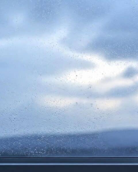 Ventana con lluvia y cielo nublado que empieza a abrirse, con una taza blanca en el alféizar, como imagen de la diferencia entre tristeza y depresión