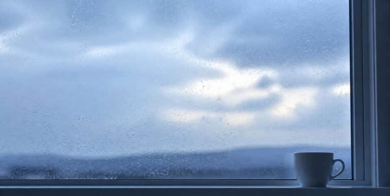 Ventana con lluvia y cielo nublado que empieza a abrirse, con una taza blanca en el alféizar, como imagen de la diferencia entre tristeza y depresión