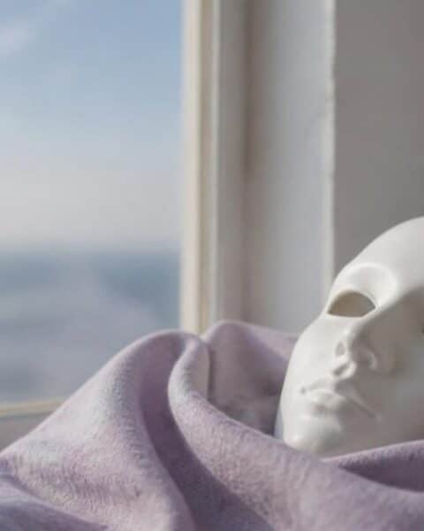 Máscara blanca de cerámica sobre una tela lila junto a una ventana