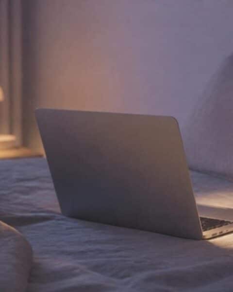 Computador abierto sobre una cama de noche, con una lámpara tenue y un vaso de agua